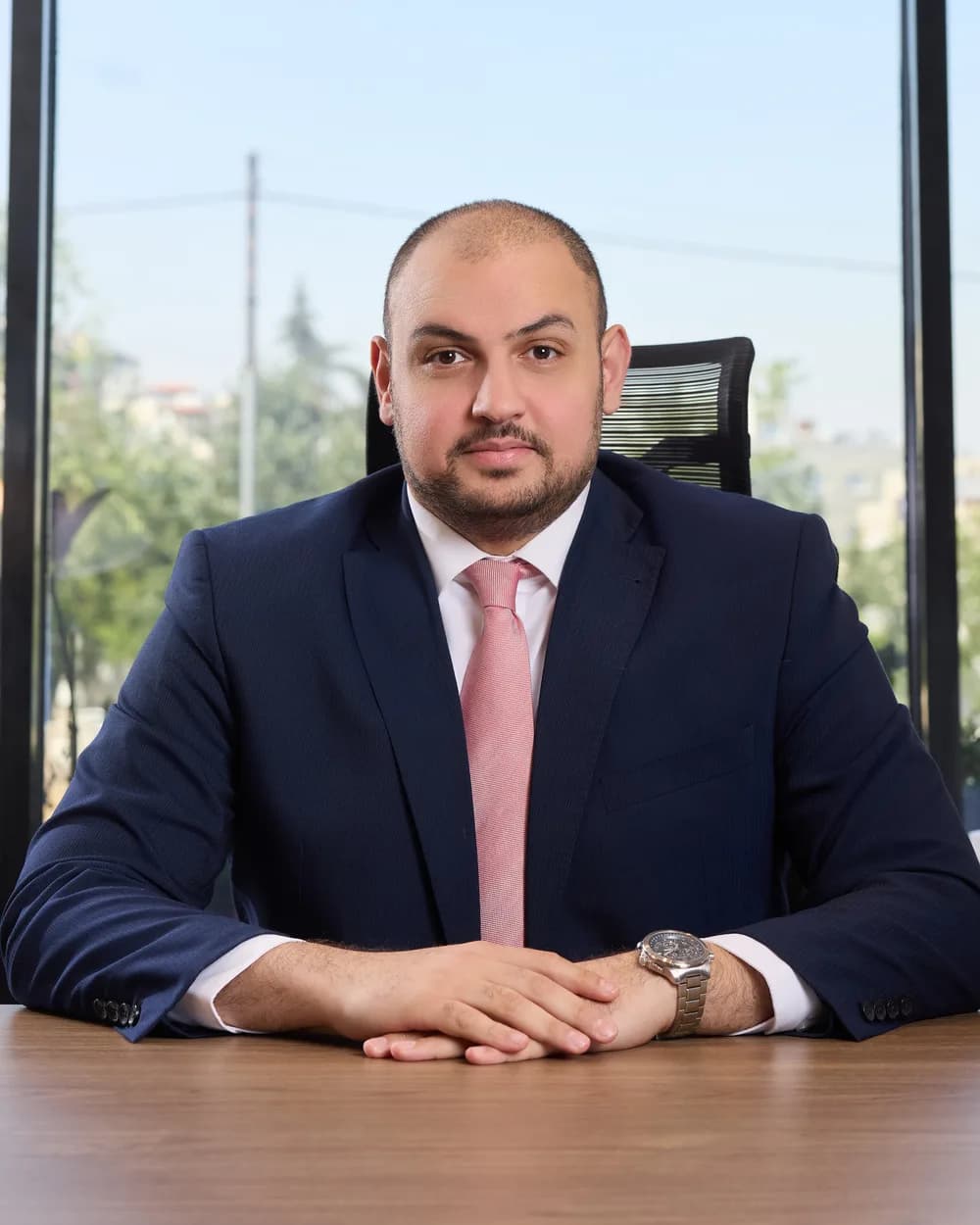 The Director of Investments of Al Kayan Hotel Investments seated at a desk in a navy suit and rose tie, hands folded, daylight and greenery behind him